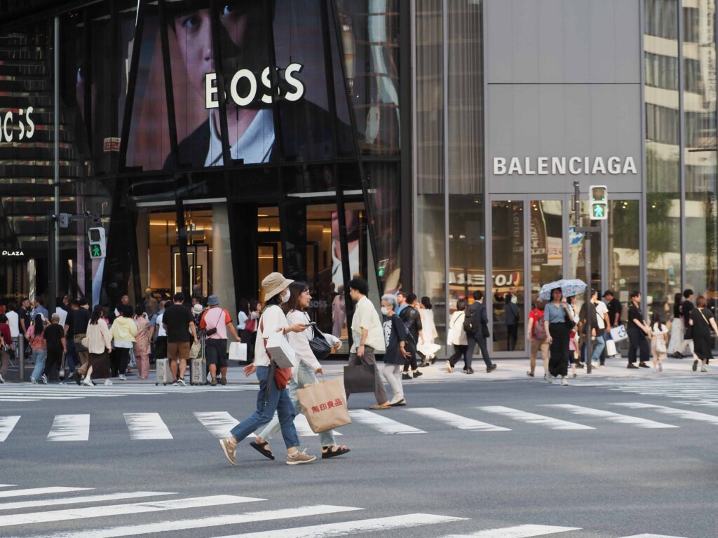Pedestrians crossing a busy city intersection in front of luxury retail stores, including Balenciaga and Hugo Boss, in an urban shopping district